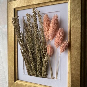 Könnte beinhalten: Ein gerahmtes gepresstes Blumenarrangement mit getrockneten rosa und braunen Gräsern in einem goldenen Rahmen.