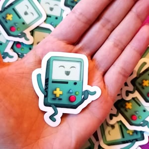 May include: A green and black cartoon character sticker of a video game console with a smiling face and arms and legs. The character is wearing a red button and has a green plus sign on the front.