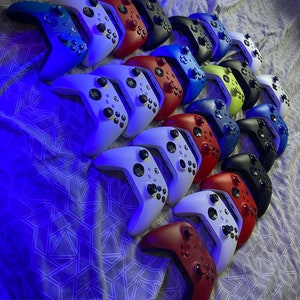 May include: A collection of 14 colourful video game controllers, mostly white, blue and red, arranged in a row on a patterned fabric surface.