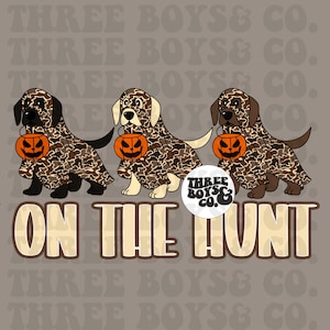 May include: Three dogs dressed in camouflage costumes holding jack-o'-lanterns. The dogs are a black lab, a yellow lab, and a brown lab. The text "THREE BOYS & CO." and "ON THE HUNT" is below the dogs.