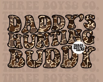 May include: A brown and tan camouflage print graphic with the text "Daddy's Hunting Buddy" and a small logo that says "Three Boys & Co."