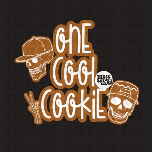 May include: White text on a brown background reads "One Cool Cookie" with a skull wearing a baseball cap on either side of the text. A small circle with white text reads "Three Boys & Co."