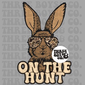 May include: A brown rabbit wearing camouflage sunglasses with the text "ON THE HUNT" in brown and white letters. The rabbit is wearing a round sticker that says "THREE BOYS & CO."