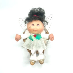 Può includere: Una piccola bambola con i capelli neri e un vestito bianco è seduta su una sedia di vimini. La bambola ha un gilet verde e bianco con un cuore rosso. La bambola ha le ali e indossa una fascia rossa.