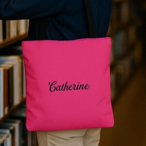 May include: A bright pink tote bag featuring the name Catherine printed in a black cursive script font. The bag has two black shoulder straps and a square shape, providing a simple and functional design for carrying various personal items.
