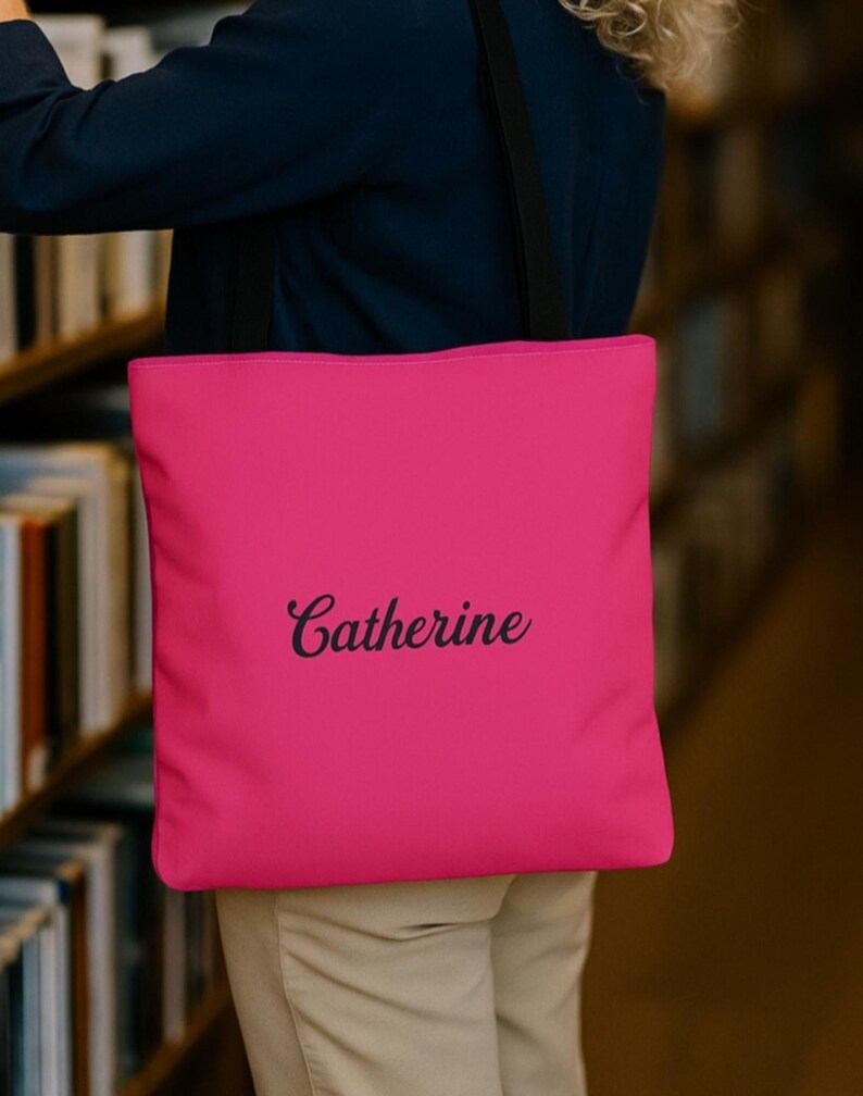 May include: A bright pink tote bag featuring the name Catherine printed in a black cursive script font. The bag has two black shoulder straps and a square shape, providing a simple and functional design for carrying various personal items.