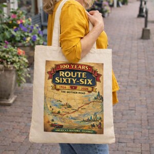 May include: A beige canvas tote bag featuring a graphic print of a Route 66 map. The design includes the text 100 YEARS ROUTE SIXTY-SIX 1926-2026 THE MOTHER ROAD, a red banner, a red vintage car, and a red dotted line tracing the historic highway route.