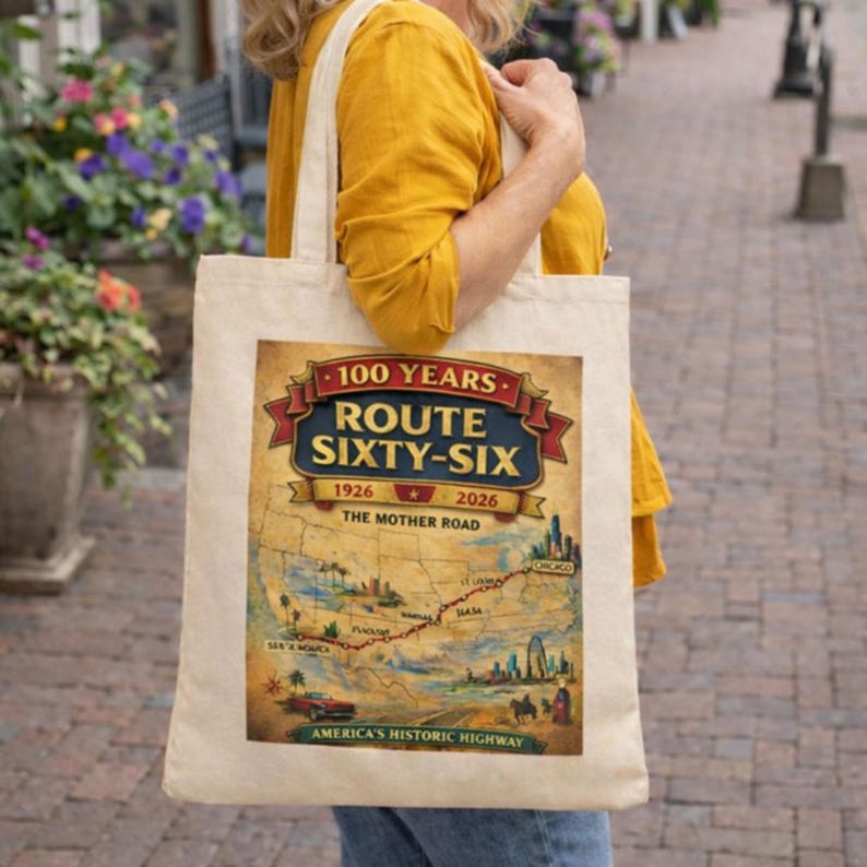 May include: A beige canvas tote bag featuring a graphic print of a Route 66 map. The design includes the text 100 YEARS ROUTE SIXTY-SIX 1926-2026 THE MOTHER ROAD, a red banner, a red vintage car, and a red dotted line tracing the historic highway route.