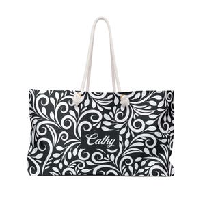 A spacious black-and-white weekender tote covered in an elegant swirling leaf and vine pattern, with white shapes forming bold, flowing designs against a black background. In the center, a black nameplate displays the name “Cathy” in graceful white script, showing that the bag can be personalized with any name. The tote has thick white rope handles threaded through gold-toned grommets at the top, giving it a sturdy and stylish look.