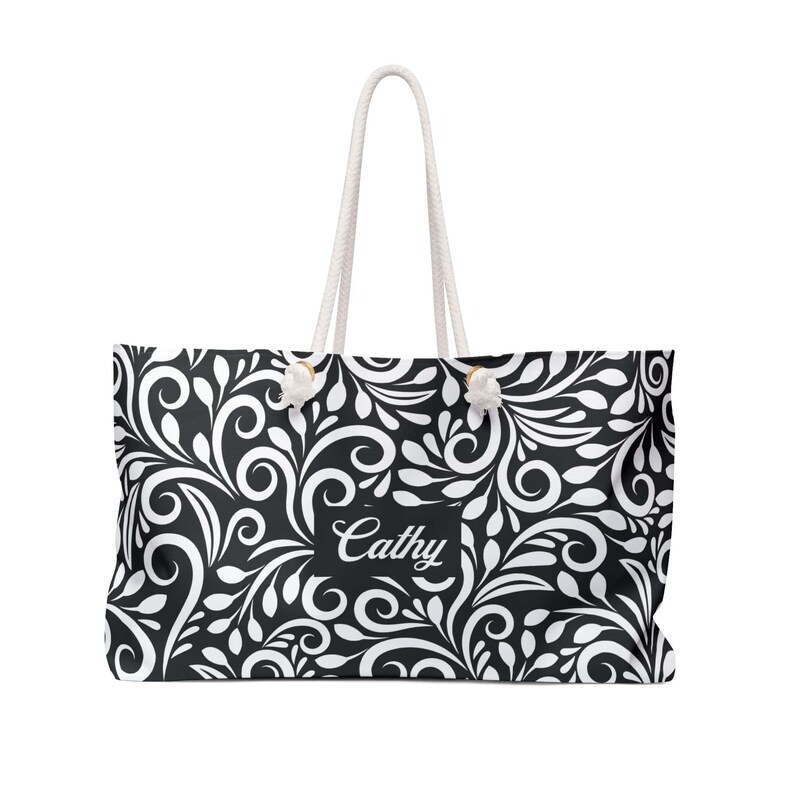 A spacious black-and-white weekender tote covered in an elegant swirling leaf and vine pattern, with white shapes forming bold, flowing designs against a black background. In the center, a black nameplate displays the name “Cathy” in graceful white script, showing that the bag can be personalized with any name. The tote has thick white rope handles threaded through gold-toned grommets at the top, giving it a sturdy and stylish look.