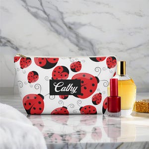 May include: A white cosmetic bag featuring a repeating pattern of red ladybugs with black spots and thin black swirls. A black rectangular label in the center displays the name Cathy in white script. A gold zipper runs along the top edge.