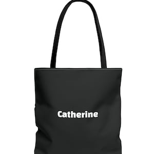 A simple, modern black polyester tote bag with clean lines and a smooth, solid-color surface. In the center, the name “Catherine” appears in bold white lettering, showing that the bag can be personalized with any name. The tote features sturdy black shoulder straps attached at the top.