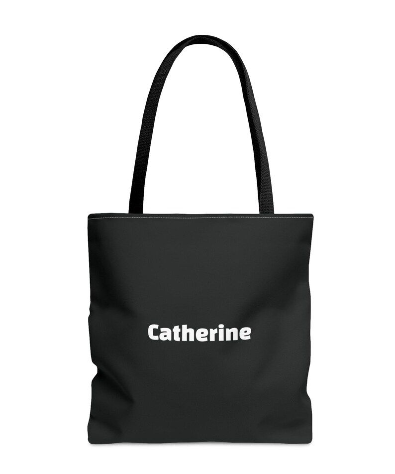 A simple, modern black polyester tote bag with clean lines and a smooth, solid-color surface. In the center, the name “Catherine” appears in bold white lettering, showing that the bag can be personalized with any name. The tote features sturdy black shoulder straps attached at the top.