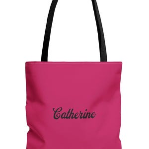 A simple, solid pink polyester tote bag with a clean and modern look. The front features the name “Catherine” printed in a smooth black script, showing that the bag can be personalized with any name. The tote has sturdy black shoulder straps that contrast against the bright pink bag. The overall design is bold, minimal, and stylish.