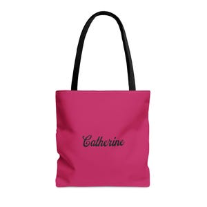 May include: A bright pink tote bag featuring the name Catherine printed in a black cursive script font in the center. The bag has two sturdy black shoulder straps attached to the top, providing a clear contrast against the vibrant pink fabric.