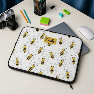 May include: White laptop sleeve featuring a repeating pattern of cartoon bees in yellow and black. The sleeve has a black zipper and a gold rectangle with the name "Cathy" printed on it. The sleeve is on top of a laptop.