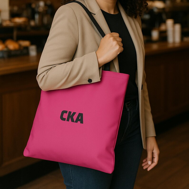 May include: A bright pink tote bag with black shoulder straps and the letters CKA printed in bold black font on the front. The bag is made of a smooth material and features a rectangular shape with a flat bottom and a spacious main compartment.