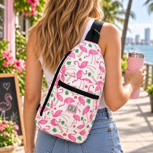 May include: A white sling backpack featuring a repeating pattern of pink flamingos and green tropical leaves. The bag has a black zip, a black strap, and a rectangular patch on the front pocket with the letters CKA printed in green.