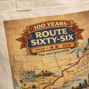 May include: A beige canvas tote bag featuring a graphic print of a map of Route 66. The text reads 100 YEARS ROUTE SIXTY-SIX 1926-2026 THE MOTHER ROAD. The map includes illustrations of city skylines and a red line tracing the route across the United States.