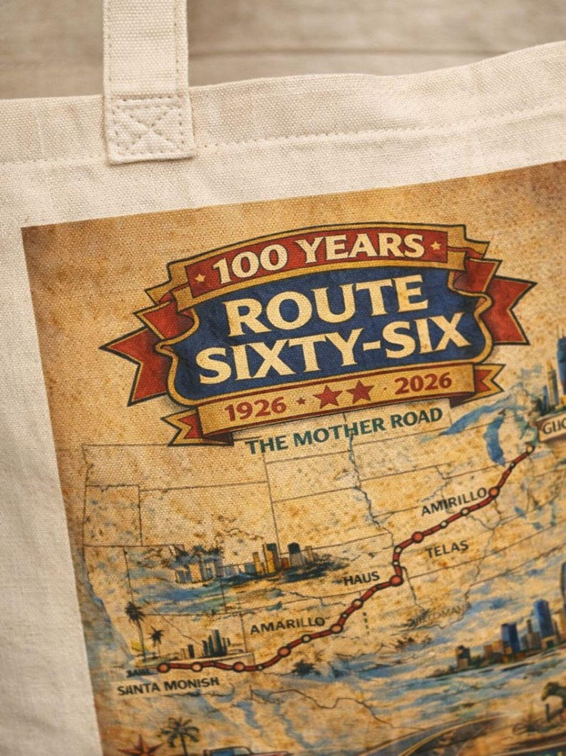 May include: A beige canvas tote bag featuring a graphic print of a map of Route 66. The text reads 100 YEARS ROUTE SIXTY-SIX 1926-2026 THE MOTHER ROAD. The map includes illustrations of city skylines and a red line tracing the route across the United States.