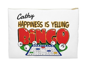 May include: A white zippered pouch featuring the text Cathy HAPPINESS IS YELLING BINGO in black, yellow, and red. The design includes a blue bingo card and several white bingo balls with numbers scattered on a green circular background.