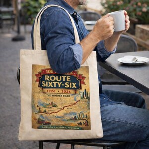 May include: A beige canvas tote bag featuring a graphic print of a map of Route 66. The design includes the text 100 YEARS ROUTE SIXTY-SIX 1926-2026 THE MOTHER ROAD AMERICA'S HISTORIC HIGHWAY, along with illustrations of a car, city skylines, and a red route line.