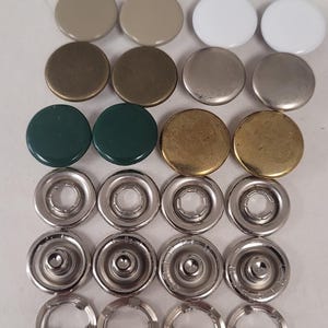 May include: A collection of circular metal snap buttons arranged in rows. The set includes smooth caps in navy, beige, white, metallic bronze, silver, dark green, and gold finishes, alongside silver-toned metal socket and stud components.