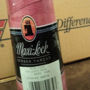 Peut inclure: Une bobine de fil à surjet Maxi-Lock Dusty Rose. L'étiquette indique 2743 mètres de fil. La bobine est enveloppée dans du plastique transparent et a un dessus blanc. En arrière-plan, une boîte en carton.