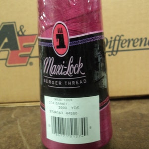 Peut inclure: Une bobine de fil à coudre sergé Maxi-Lock rose avec une étiquette blanche indiquant "Maxi-Lock Serger Thread", "274 Garnet", "3000 YDS", "9724143 44586" et un code-barres.