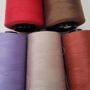 May include: Five spools of thread in different colors: red, brown, purple, beige, and orange. The spools are arranged in a semi-circle on a white surface.
