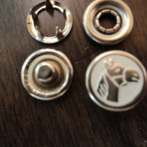 May include: Four silver metal snap fasteners, two with a white background and a silver horse head design, one with a silver ring and three prongs, and one with a silver ring and a silver center.