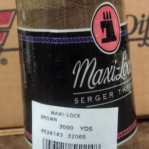 Peut inclure: Une bobine de fil à coudre pour surjeteuse Maxi-Lock de couleur marron avec une étiquette blanche indiquant "Maxi-Lock Serger Thread, Brown, 3000 YDS, 4524143 32065" et un code-barres.