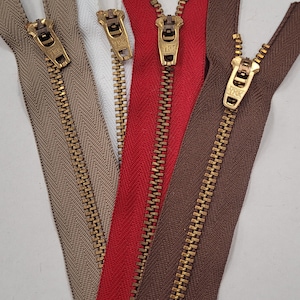 May include: Five different colored zippers with gold metal teeth and pulls. The zippers are brown, beige, white, red, and dark brown.