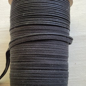 May include: A reel of black elastic cord. The cord is tightly wound around a cardboard reel. The elastic appears to be made of a woven material. The reel is sitting on a white surface.