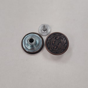 May include: A set of three metal jean buttons. One button is silver with a raised center, and the other two are copper with a decorative floral design.