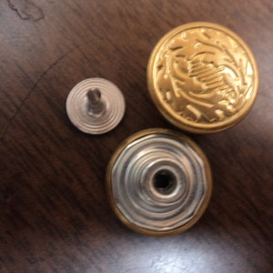 May include: Three metal buttons, two gold and one silver. The gold buttons have a raised design and the silver button has a concentric circle design.
