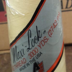 Peut inclure: Une bobine de fil à coudre Maxi-Lock de couleur crème. La bobine est étiquetée "Maxi-Lock Fil à coudre 3000 YDS. (2742 m) Fabriqué aux États-Unis par American & Efird Inc."