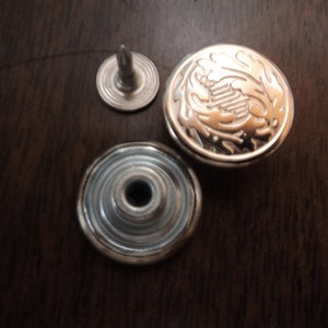 May include: A set of three silver-toned metal buttons with a decorative design. The top button has a raised floral pattern, the middle button is a flat disc with concentric circles, and the bottom button is a small stud with a pointed top.