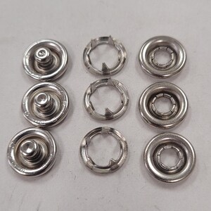May include: A collection of silver-colored snap fasteners, arranged in three rows. The fasteners include the male and female components, designed for securing fabric or other materials. The components are made of metal.