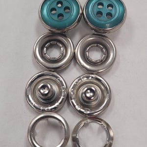 May include: A set of teal colored snap buttons with silver metal settings. The set includes two top buttons, two bottom buttons, and two caps.