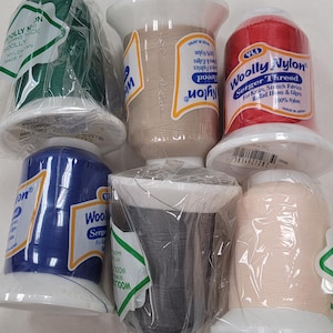 May include: An assortment of spools of Woolly Nylon serger thread in various colors, including green, tan, red, blue, gray, and peach. Each spool is wrapped in clear plastic and has a white base. The thread is labeled for use with knit and stretch fabrics.