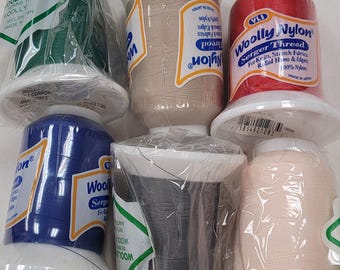 May include: An assortment of spools of Woolly Nylon serger thread in various colors, including green, tan, red, blue, gray, and peach. Each spool is wrapped in clear plastic and has a white base. The thread is labeled for use with knit and stretch fabrics.