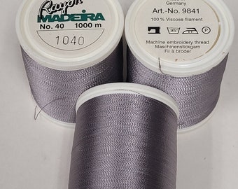May include: Three spools of gray rayon embroidery thread, each with a white label that reads "Rayon Madeira No. 40 1000 m 1040". The label also includes a sewing machine icon, a 95 degree symbol, an iron icon, and a "P" symbol. The label also reads "Made in Germany Art.-No. 9841 100% Viscose filament Machine embroidery thread Maschinenstickgarn Fil à broder".