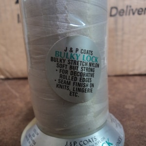 May include: A spool of white thread labeled "J & P Coats Bulky Lock Bulky Stretch Nylon" with the text "Soft But Strong" and "For Decorative Rolled Edges Seam Finish On Knits, Lingerie Etc."
