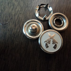 May include: Four silver metal snap fasteners, one with a white background and a design of two crossed pistols.