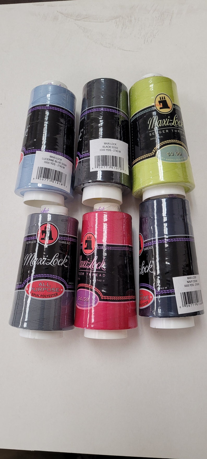 May include: Six spools of Maxi-Lock serger thread in various colors, including blue, black, green, gray, red, and navy. Each spool is labeled with the color name, thread type, and yardage. The spools are arranged in two rows of three.