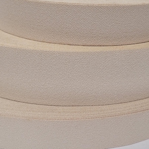 May include: Three rolls of beige elastic fabric, stacked on top of each other. The fabric is woven with a subtle texture.