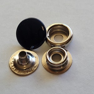 May include: A set of four black and silver snap fasteners. The fasteners are made of metal and have a round shape. The black fastener is a cap, and the silver fasteners are a socket, a stud, and a base.
