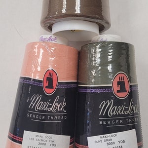 May include: Three spools of Maxi-Lock serger thread. The spools are brown, salmon pink, and olive drab. Each spool has a label with the Maxi-Lock logo, the color name, and the yardage. The labels also include barcodes and product numbers.