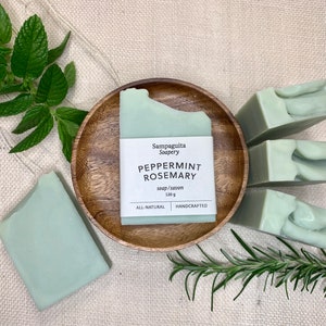 May include: Four bars of handmade peppermint rosemary soap, each bar is a light green colour and has a swirl pattern. The soap is displayed on a wooden plate with a label that reads "Sampaguita Soapery Peppermint Rosemary soap/savon 120g All Natural Handcrafted". There are sprigs of rosemary and mint leaves in the background.